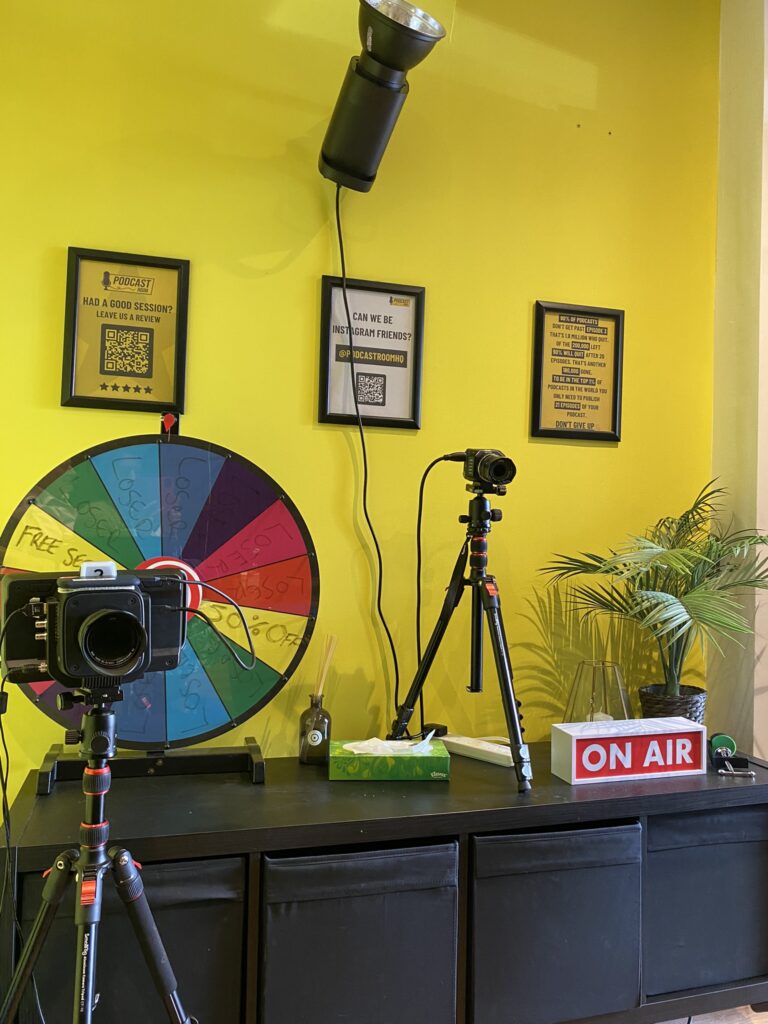 This image shows a brightly lit podcast recording setup. The background wall is painted a vibrant yellow and features three framed signs. In the foreground, there are two tripods, each holding a camera, positioned on top of a black storage unit. A colorful spin wheel labeled with different segments is also placed on the storage unit. Other items on the unit include a box of tissues, a small decorative plant, and a rectangular "ON AIR" sign. A black spotlight is mounted on the wall above the setup, casting light downward. The overall scene is organised, colourful, and clearly intended for recording video content, for a podcast or a live streaming session.